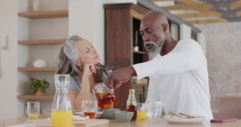 Interracial senior couple sharing relaxed breakfast at kitchen island, pouring tea