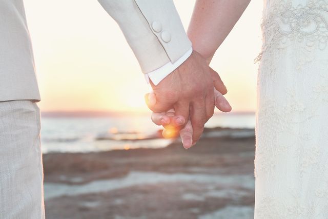The image captures a close up of a bride and groom holding hands on a beach at sunset. The soft hues and warm glow create a romantic and intimate atmosphere. Ideal for use in wedding invitations, romance novels, engagement announcements, and blogs about beach weddings or relationship advice.