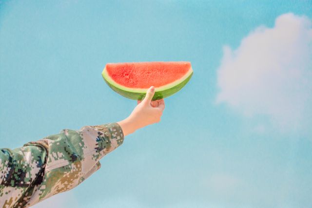 Hand holding a juicy watermelon slice against a clear blue sky with a few white clouds. Person wearing a camouflage jacket. Perfect for summer promotions, healthy eating campaigns, outdoor activities marketing, and fresh fruit advertisements.
