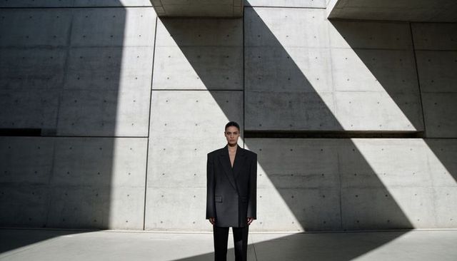 Model wearing oversized black suit in minimalist concrete atrium under angular shadows