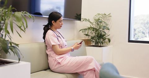 Nurse in pink scrubs checking smartphone in clinic lounge with stethoscope and smartwatch