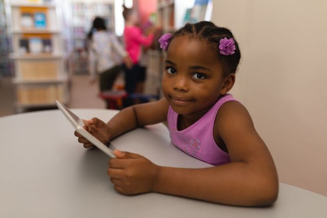 Young African American schoolgirl using digital tablet in a library, smiling at camera. Ideal for educational content, technology in education, childhood learning, and school-related materials.