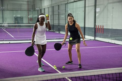 Two women training on purple padel court with paddles and yellow balls, diverse athletes