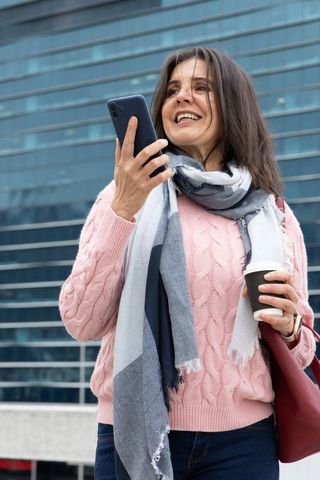 Middle-aged woman smiling while talking on phone holding coffee outside glass office building