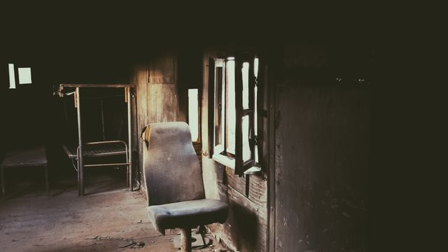 Dimly lit, abandoned room featuring dusty office chair and old bed frame. Suitable for themes of neglect, decay, solitude, or historical settings.