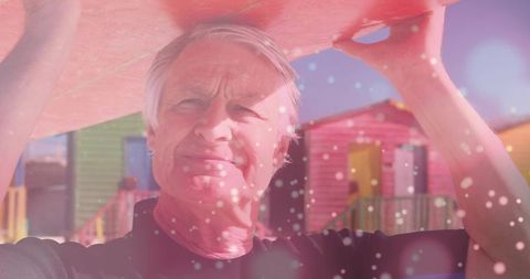 Senior man holding pink surfboard above head near colorful beach huts with sunlit bokeh