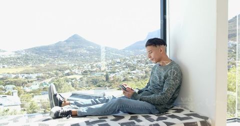Young man scrolling phone on bright window seat with panoramic mountain town view