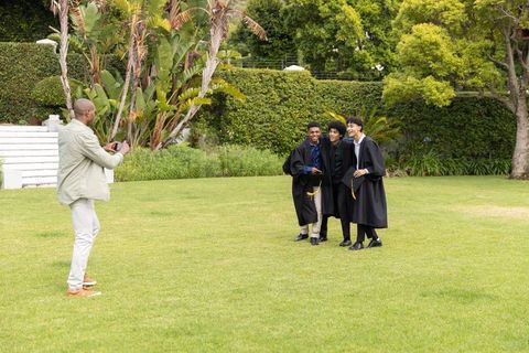 Three male graduates posing on lawn holding caps and tassels while friend shoots with phone