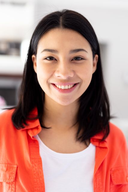 Vertical portrait of happy biracial woman in orange shirt smiling to camera at home, with copy space. Health, happiness and domestic life concept.