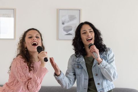 Female friends singing duet with handheld microphones in cozy living room karaoke session