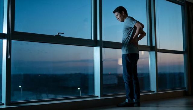 Asian man standing at high-rise window at dusk holding lower back contemplating city lights
