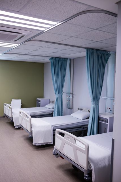 Image depicts an empty hospital ward with multiple beds, each separated by blue curtains. The setting is clean and sterile, ideal for patient care and recovery. This image can be used for healthcare-related content, medical facility promotions, or articles discussing hospital environments and patient care.