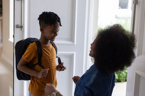 Mother greeting smiling son carrying backpack at home entrance for school morning routine