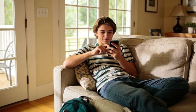 Teen relaxing on sofa scrolling smartphone by sunlit french doors with backpack