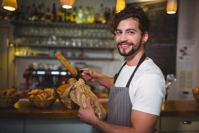 Young waiter in a cafe packing fresh croissants into a paper bag while smiling at the camera. Ideal for use in content related to food service, hospitality, small businesses, cafes, and customer service. Perfect for illustrating friendly and welcoming service in a bakery or cafe setting.