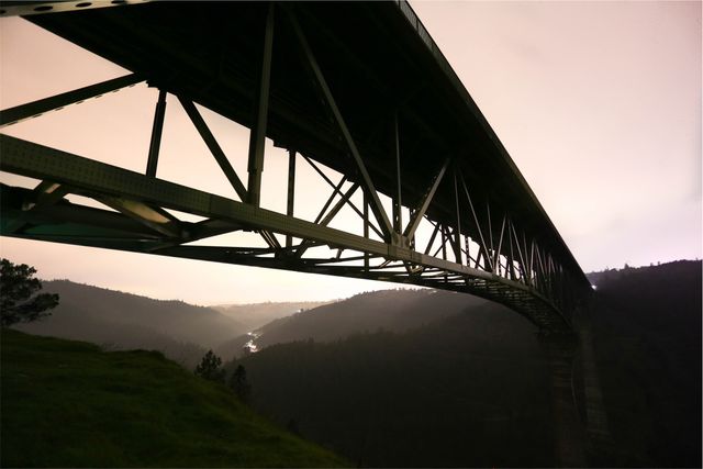 Spanning across towering mountainous slopes, an iron bridge appears silhouetted against the twilight sky. Below, the serene valley stretches into the distance. Ideal for use in projects related to engineering marvels, infrastructure development, transportation systems, travel documentaries, scenic calendars, architectural presentations, and enhancing articles on adventure and exploration.