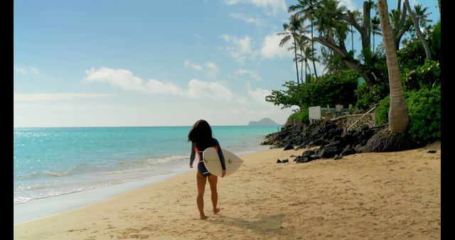 A woman walks on a tropical beach carrying a surfboard under clear blue sky. Perfect for promoting beach vacations, surf camps, travel destinations, and outdoor activities. Also ideal for articles about adventure sports, leisure, and holiday experiences.