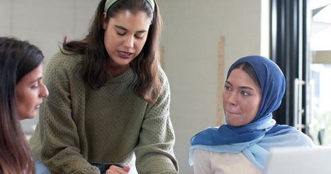 Diverse female coworkers collaborating at desk woman wearing blue hijab reviewing documents