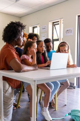 African American teacher guiding diverse children collaborating around laptop in classroom