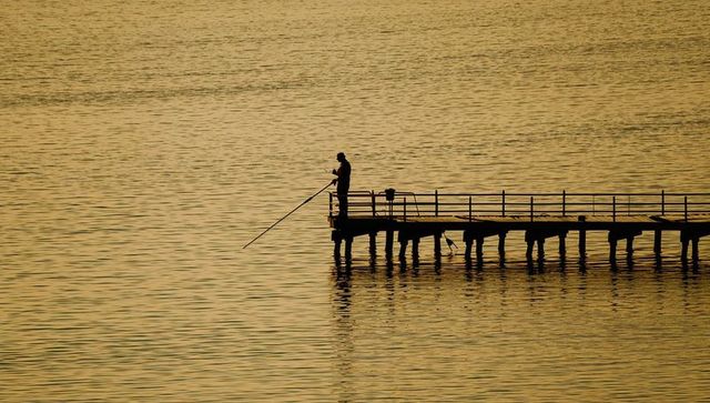 Silhouetted angler fishing on wooden pier during golden hour sunset with water reflection