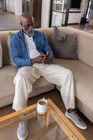 Senior african american man sitting on sofa using smartphone in modern living room