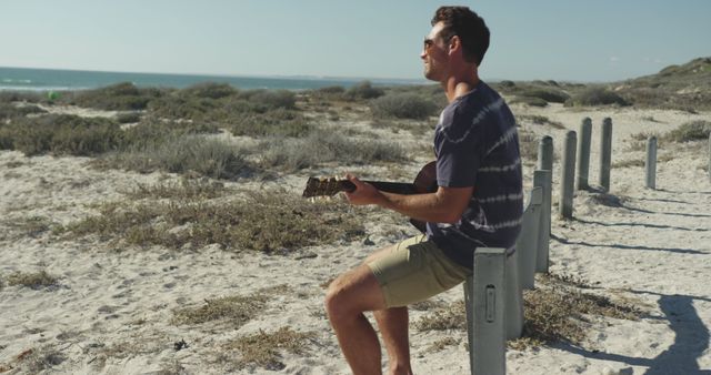 Happy caucasian man sitting on beach by the sea playing guitar. beach stop off on summer holiday road trip.