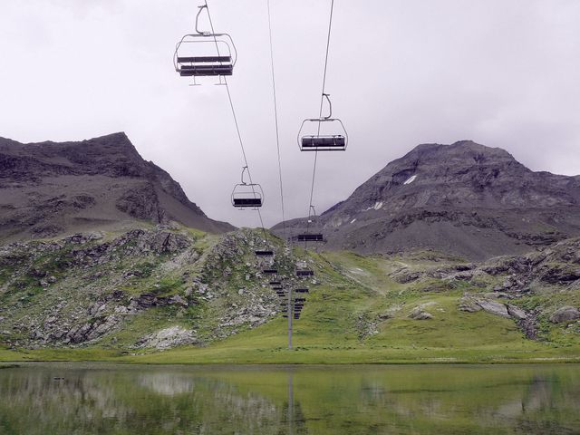 A row of empty cable cars extending over a serene mountain lake under overcast skies. Rugged mountains and lush green terrain create a peaceful and natural scene, perfect for promoting adventure travel, outdoor activities, and nature tourism.