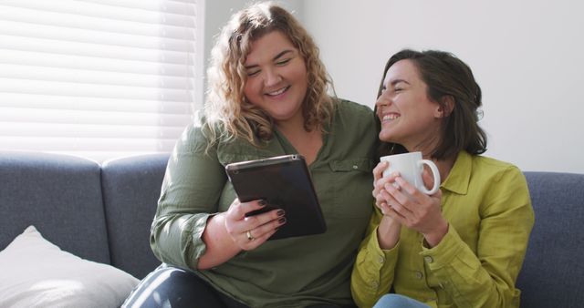 Caucasian lesbian couple using tablet, drinking coffee and sitting on couch. domestic life, spending free time relaxing at home.