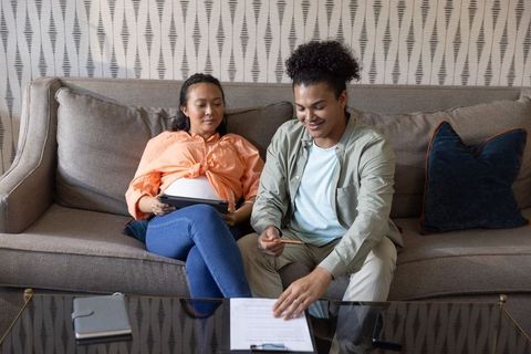 Couple completing home paperwork while expecting baby, pregnant woman using tablet on sofa