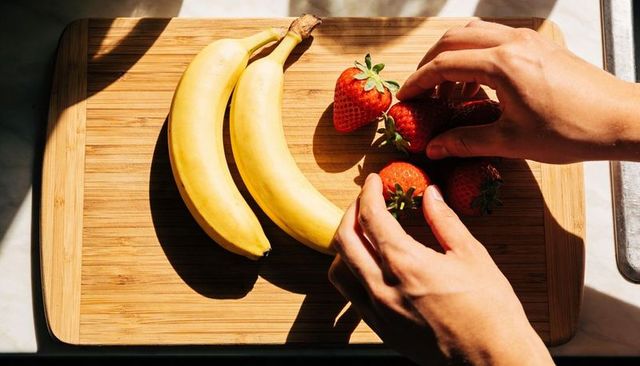 Arranging strawberries and bananas on wooden cutting board in sunlight with hands