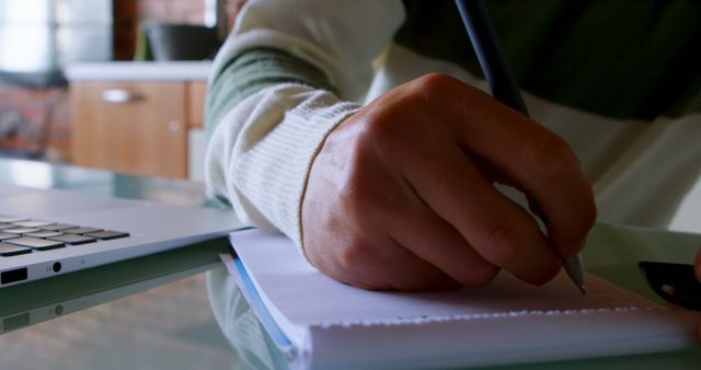 This close-up image shows a person writing in a notebook next to a laptop on a desk, making it ideal for illustrating concepts related to studying, planning, working from home, or busy office environments. It can be used in blogs, articles, and promotions concerning education, productivity, or digital workspaces.