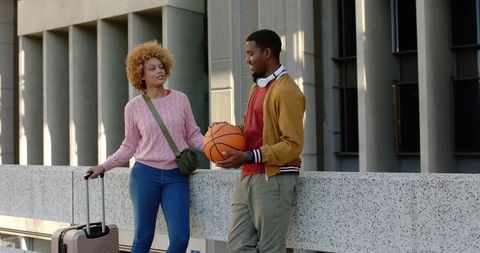 Young couple chatting and leaning on urban terrace while carrying suitcase and basketball