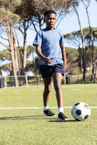 African american boy dribbling soccer ball on sunny turf field with pine trees background