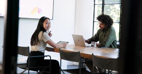 Asian woman and Black man collaborating over pie chart with tablet and laptop