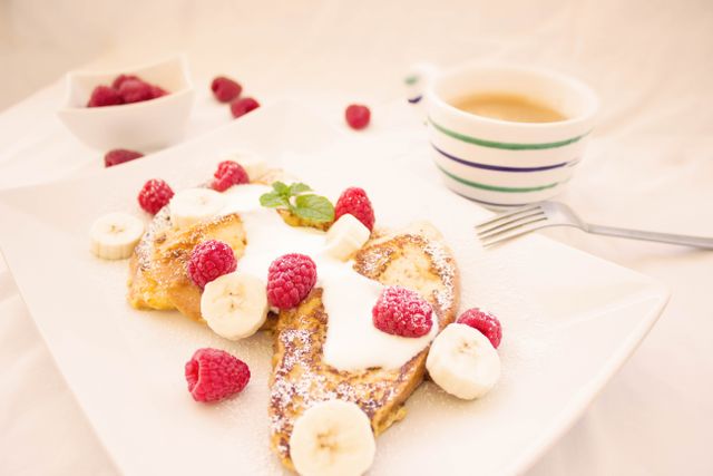 French toast topped with fresh raspberries, banana slices, powdered sugar and yogurt served on a white plate, with a cup of coffee and a bowl of raspberries next to it. This image is perfect for food blogs, breakfast menus, healthy eating websites, and social media posts highlighting breakfast ideas.