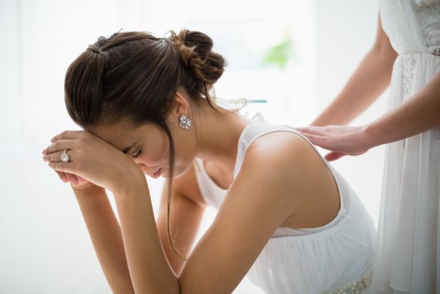 This image captures an emotional moment of a bride sitting with her hand on her forehead, possibly feeling overwhelmed or stressed. A supportive friend or bridesmaid is seen comforting her by placing a hand on her shoulder. This image can be used in articles or blogs about wedding planning, emotional support during weddings, bridal stress, or the emotional aspects of wedding preparations.