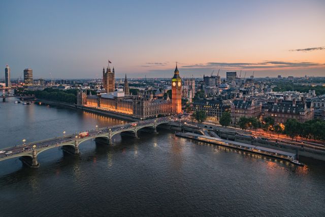 Stunning aerial view of the Houses of Parliament and Big Ben at sunset, with the Thames River and bridge creating picturesque scenery. Ideal for use in travel brochures, websites promoting London tourism, educational materials about British landmarks, or evening cityscapes.