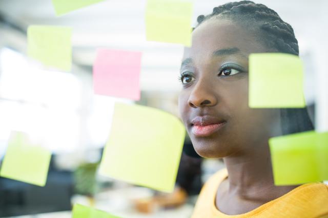 African American female graphic designer analyzing colorful sticky notes on glass in modern office. Ideal for content related to creativity, business planning, brainstorming sessions, professional work environments, and strategic thinking.