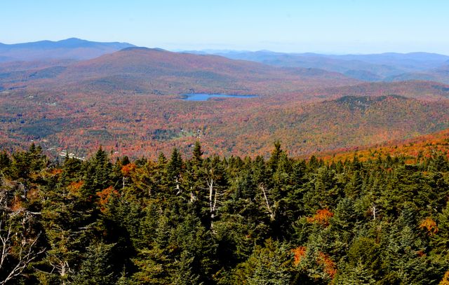 This view highlights a stunning autumn landscape with vibrant fall foliage covering a vast mountain range. A scenic lake sits serenely amid the dense trees, creating a peaceful atmosphere. Perfect for advertising nature retreats, outdoor activities, seasonal tourism, travel promotions, and calendars focused on natural beauty.