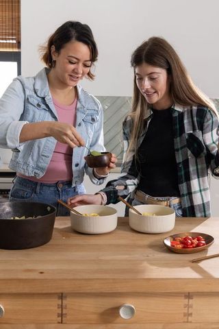 Two women preparing pasta and garnishing bowls at wooden kitchen island in home kitchen