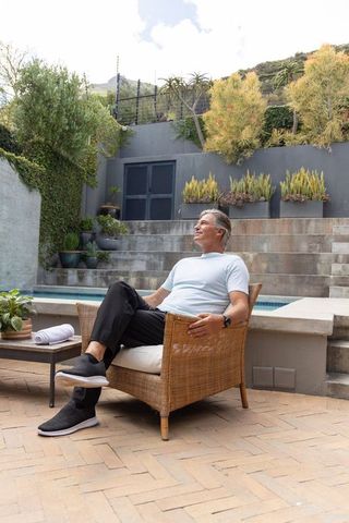 Mature man relaxing on patio chair beside pool checking smartwatch in landscaped backyard