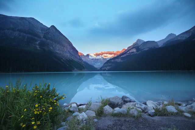 Sunrise illuminates mountain peaks surrounding a calm lake with clear reflections. Yellow wildflowers bloom by rocky shore in foreground. Ideal for nature-themed designs, travel brochures, adventure promotions, or calming social media content, this image captures serene beauty and tranquility of untouched wilderness.