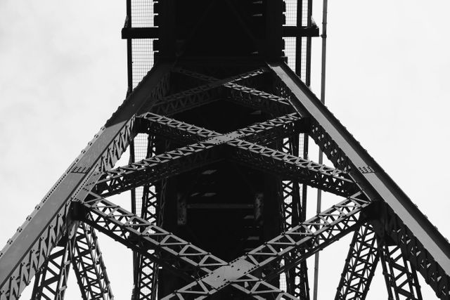 Capturing the under-structure of an iron bridge in black and white, this image highlights the intersecting metal beams and detailed geometric patterns. Its architectural design and industrial elements can be used for design projects, promotional materials for construction companies, educational purposes, or art pieces emphasizing structural beauty and engineering.