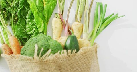 Burlap sack holding fresh organic vegetables in minimal flat lay on white surface, rustic produce