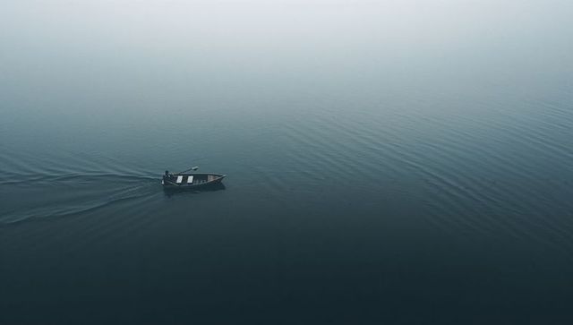 Solitary rower crossing misty lake in wooden rowboat, minimalist calm and solitude