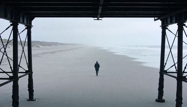 Solitary figure walking under pier on misty beach leaving footprints to horizon
