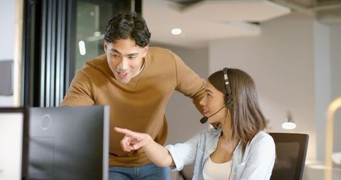 Indian woman and coworker collaborating at workstation, headset pointing at monitor