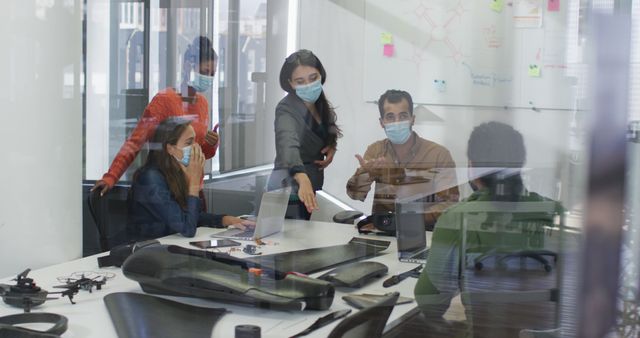 People are collaborating and brainstorming ideas around a conference table, emphasizing workspace safety protocols by wearing PPE masks. Ideal for demonstrating modern office environment concepts, teamwork, company strategies, COVID-19 precautions, and business planning.