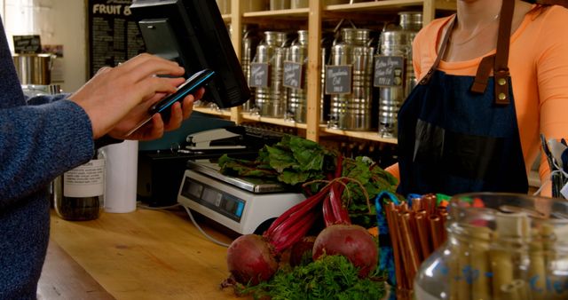 Customer using mobile payment at checkout counter in an organic grocery store. Fresh produce including beetroots and green vegetables on counter. Ideal for illustrating digital payment methods, shopping, organic food markets, and modern technology in retail.