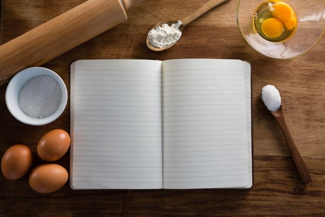 Over head view of book, eggs, flour, spoon and rolling pin kept on a table
